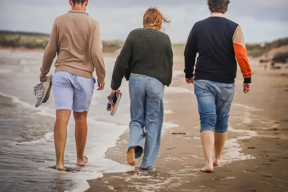 Three students walking barefoot on the beach with their backs towards the camera. Photo.