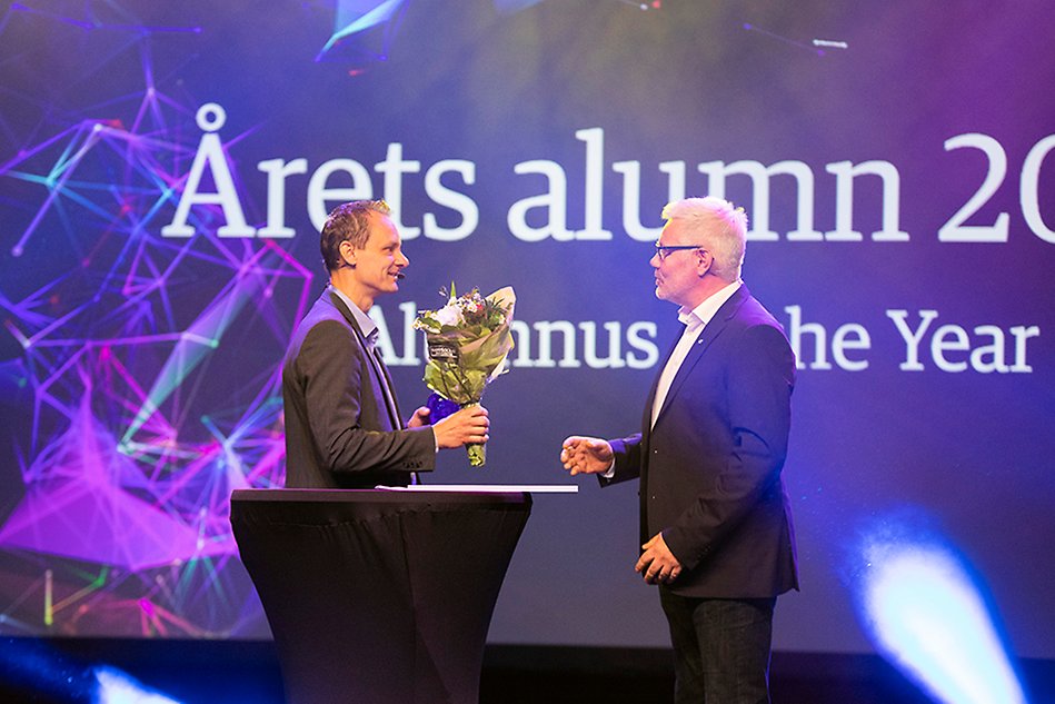 Two people visible from the side are facing each other in front of a large, colorful screen. The person on the left is holding a bouquet of flowers and the person on the right is saying something. Photo. 