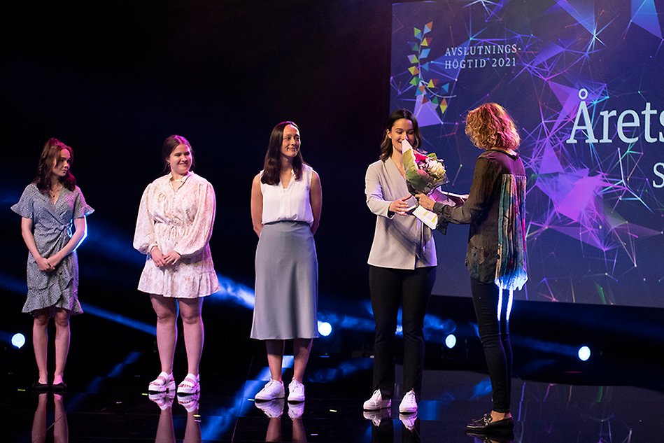 Four people stand in a row on a stage where the fourth person on the far right receives flowers from a person standing with her back to the camera. Photo.