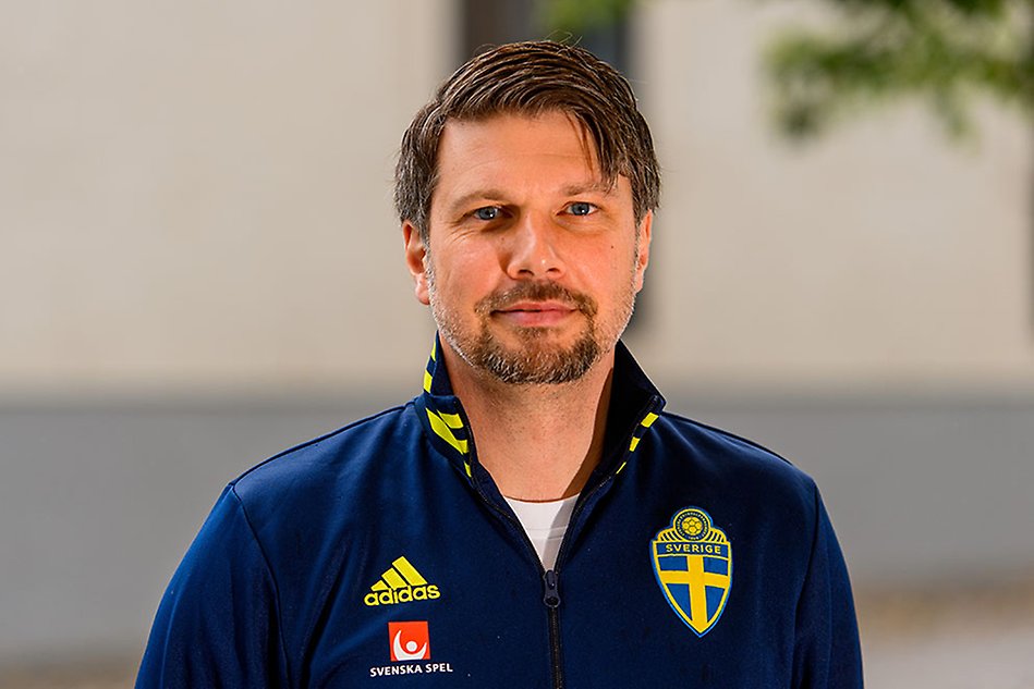 A man smiling, looking in to the camera. He is wearing a dark blue shirt with a Swedish flag on it. Photo