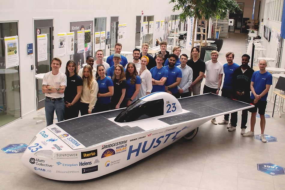 A group of students posing around a white-blue car. Photo.