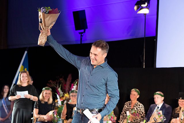 A person in a shirt and jeans holds up a bouquet of flowers in the air in a gesture of victory. Photo