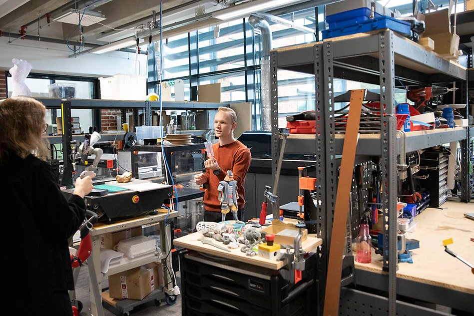 A man in an orange sweater is being filmed by a woman in dark clothes in a lab environment. Photo.