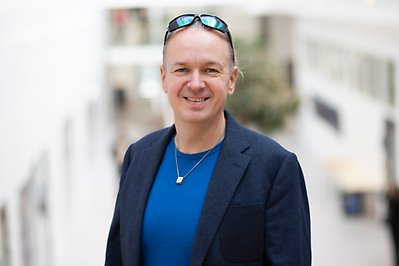 Person in a blue sweater and blue jacket with sunglasses on his forehead smiles and looks into the camera. Photo