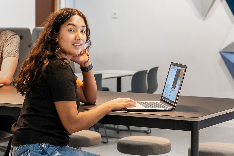 A young women is sitting at a table with a laptop infront of her. The picture is taken from the side. Photo.