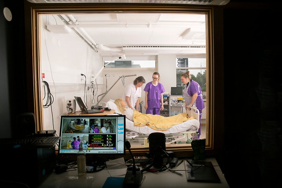En lärare instruerar två sjuksköterskestudenter vid en säng med en docka. Bilden är tagen från ett observationsrum med en bildskärm. Foto.